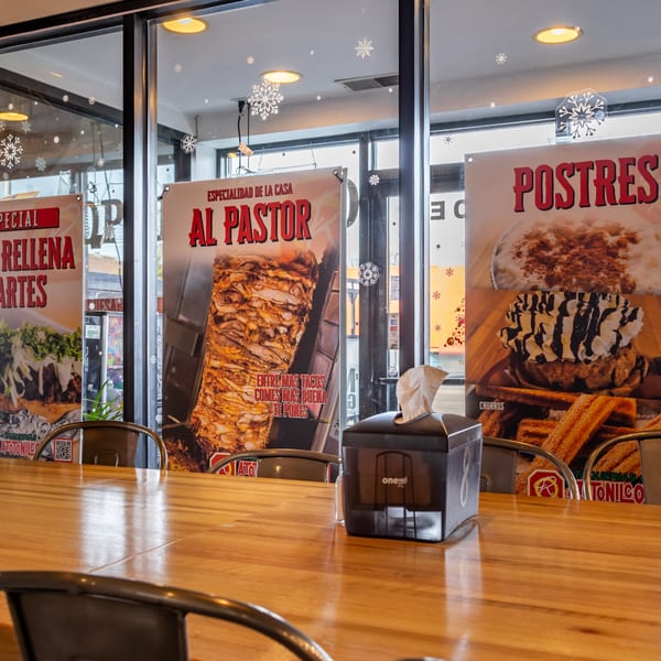 Restaurant interior with wooden tables, chairs, and large window posters advertising Mexican food and desserts.