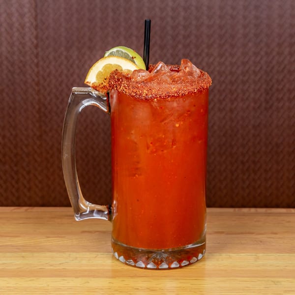 A large glass mug of red michelada with a chili-salted rim, garnished with orange and lime slices.