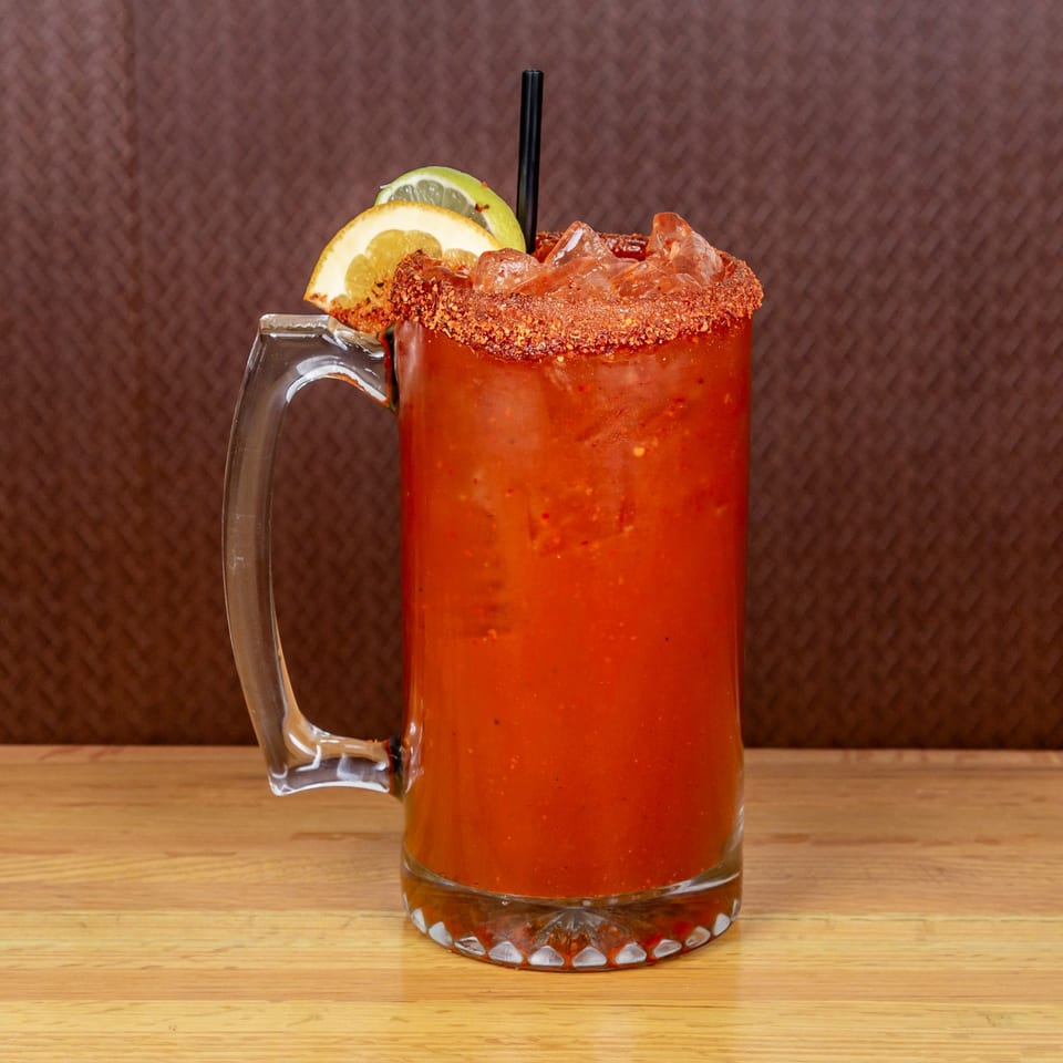 A large glass mug of red michelada with a chili-salted rim, garnished with orange and lime slices.