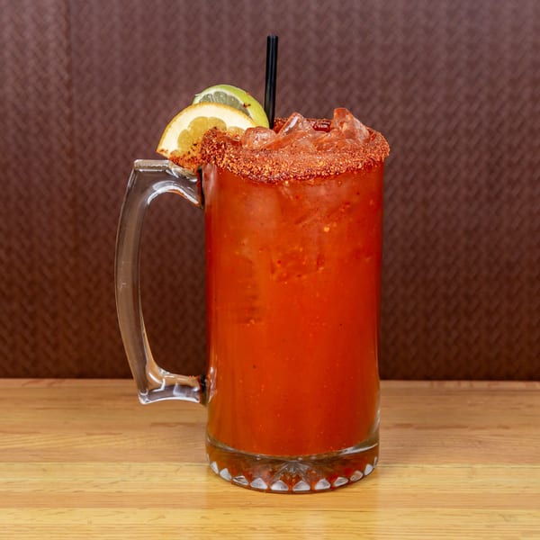 A large glass mug of red michelada, garnished with lime and orange slices, on a wooden table.