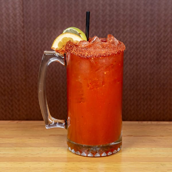 A large glass mug of red michelada with ice, a chili-lime rim, and citrus slices.