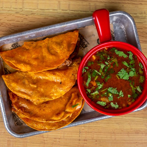 Three golden-brown birria tacos and a red bowl of consommé with cilantro on a metal tray.