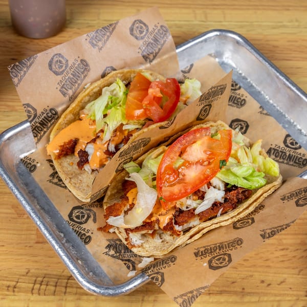 Two Taquerias Atotonilco tacos with meat, lettuce, tomatoes, and sauce on branded paper in a metal tray.
