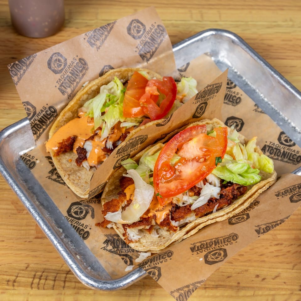Two Taquerias Atotonilco tacos with meat, lettuce, tomatoes, and sauce on branded paper in a metal tray.