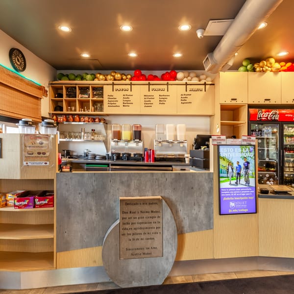 A brightly lit Mexican restaurant counter with drink dispensers, a menu, and a Coca-Cola fridge.