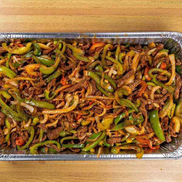 Close-up of a large foil tray filled with sizzling beef fajitas, green peppers, and onions.