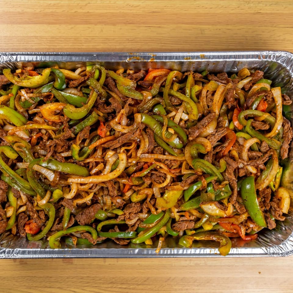 Close-up of a large foil tray filled with sizzling beef fajitas, green peppers, and onions.