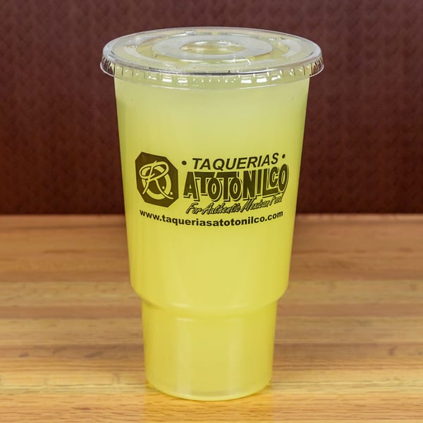 A clear plastic cup filled with a light yellow beverage, branded with Taquerias Atotonilco logo.